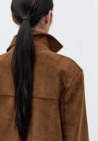 Brązowa kurtka zamszowa z wysokim kołnierzem, widocznym przeszyciem i gładką fakturą. Włosy związane do tyłu, odsłaniające tył szyi.