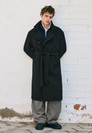 Un jeune homme pose devant un mur de briques blanches, vêtu d’un manteau noir ceinturé, d’un pantalon gris à carreaux et de chaussures foncées.