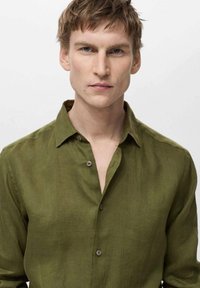 Camicia in lino verde oliva con colletto button-down, caratterizzata da un fit rilassato e una texture sottile. Il tessuto appare leggero e traspirante.