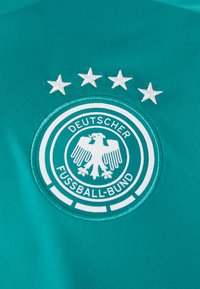 Τιρκουάζ φανέλα με κεντητό κυκλικό σήμα. Το σήμα περιλαμβάνει μια μαύρη αετό και το κείμενο "DEUTSCHER FUSSBALL-BUND", περιτριγυρισμένο από πέντε λευκά αστέρια.