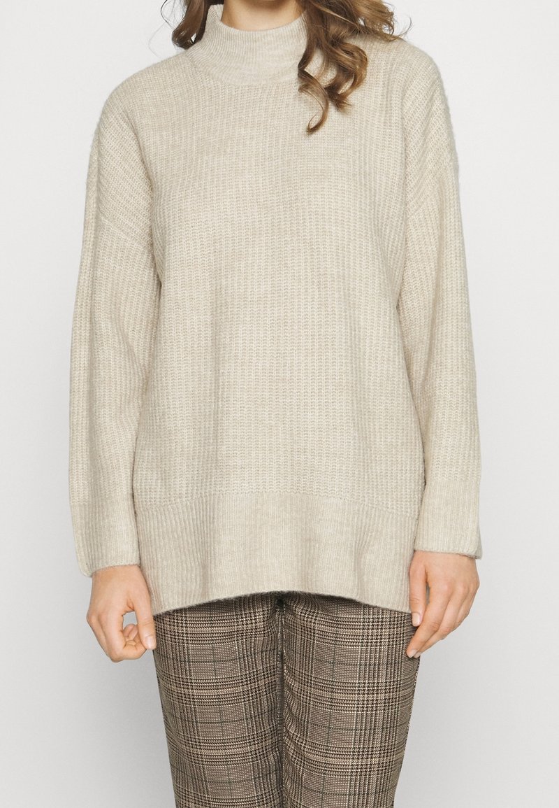 Femme portant un pull beige à col montant en maille et un pantalon à carreaux marron, debout devant un fond blanc uni.