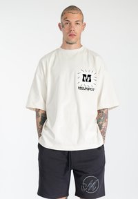T-shirt oversize crema a maniche corte con design grafico nero sulla tasca. Abbinato a pantaloni corti neri con logo "M" ricamato.