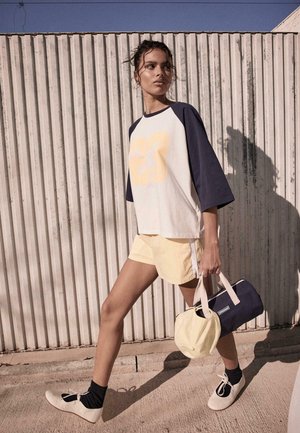 Žena kráča vonku, oblečená v bielom a námorníckom tričku s číslom 23, bledé žlté šortky, čierne ponožky a biele topánky, nesie námornícku a béžovú tašku.