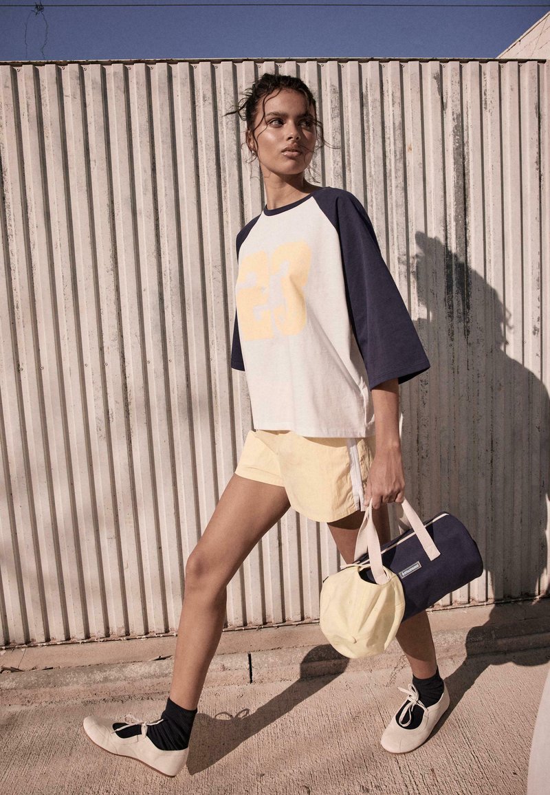 Femme marchant à l'extérieur portant un t-shirt blanc et marine avec le numéro 23, un short jaune pâle, des chaussettes noires et des chaussures blanches, portant un sac marine et beige.