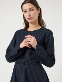 Γυναίκα που φοράει ένα φορεματάκι με ναυτικό μπλε ρίγες, μακριά μανίκια και αντίστοιχη ζώνη, ποζάροντας με τα χέρια κλεισμένα κοντά στο στήθος μπροστά σε ένα απλό φόντο.