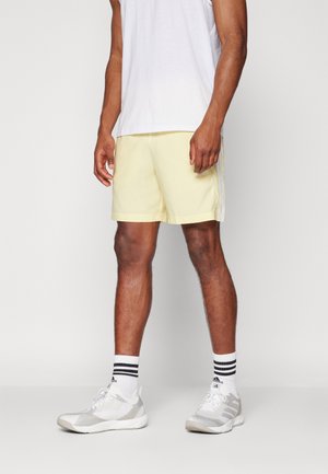 Shorts de sport jaune clair avec une texture lisse, dotés de rayures blanches sur les côtés et d'un petit logo. Associés à des baskets blanches et grises et des chaussettes rayées.