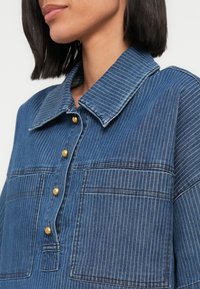 Chemise en denim avec des rayures verticales bleues et blanches, un col, une patte de boutonnage avec des boutons dorés, et deux poches poitrine.