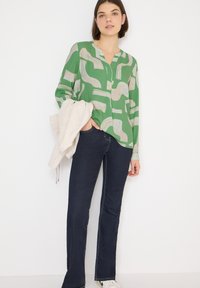 Zielona koszula z abstrakcyjnymi beżowymi wzorami, z pojedynczym guzikiem przy szyi, luźnego kroju. Połączona z ciemnymi dzwonowatymi dżinsami i jasnym swetrem zarzuconym na ramię.