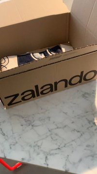 Zalando kartondoboz, barna, fekete logóval. Részben nyitva, benne egy mintás és sötét színű hajtogatott tárgy látható.