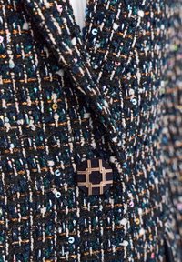 Navy tweed jakk, millel on erevärvilised sädelevad detailid ja musta ja kulla kontrastne kinnitus kaela lähedal, omades tekstureeritud kudumist.