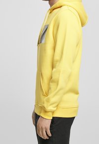 Žlutá mikina s kapucí a přední kapsou, zdobená grafickým designem na hrudi. Měkký bavlněný materiál s žebrovanými manžetami a pasem.