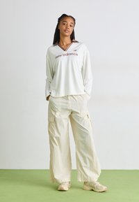 Long-sleeve, white mesh top with brown collar and “new balance” logo; cream cargo pants with pockets; beige sneakers; green floor.