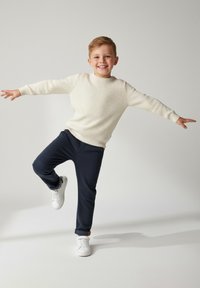 Jaśniutki beżowy prążkowany sweter, ciemnogranatowe spodnie, białe trampki; dziecko pozujące z podniesioną jedną nogą, wyciągniętymi ramionami i uśmiechnięte.