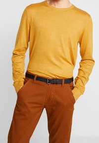 Senapsgul långärmad tröja med rund hals, tillsammans med bruna byxor och ett svart bälte med spänne. Slät, lätt material.