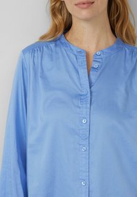 Camicia azzurra chiaro con colletto rotondo, maniche lunghe e texture morbida e liscia. Presenta piccoli bottoni lungo la parte anteriore e tessuto arricciato alle spalle.