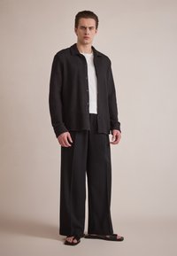 Cardigan noir en tricot avec un col, fermeture à boutons, associé à un pantalon large noir et des sandales noires sur un fond gris clair.