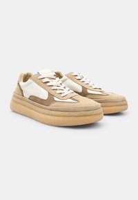 Beige velúr sportcipők fehér bőrkiegészítőkkel, texturált gumi talppal, kerek orral és lapos fűzőkkel. A talpon cikkcakk varrás található.