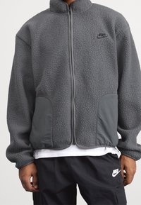 Giacca in fleece grigio Nike con colletto alto, chiusura con zip, corpo testurizzato e pannelli delle tasche grigi solidi con dettagli del logo.
