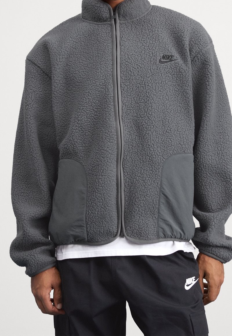 Giacca in fleece grigio Nike con colletto alto, chiusura con zip, corpo testurizzato e pannelli delle tasche grigi solidi con dettagli del logo.