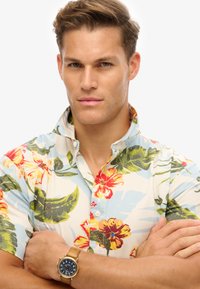 Camisa de manga corta, abotonada, con un fondo azul claro y vibrantes patrones florales en rojo, amarillo y verde; combinada con un reloj marrón.