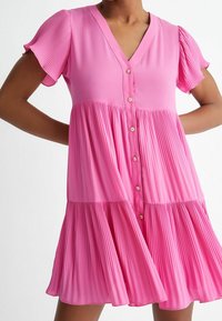 Vestido rosa plisado con escote en V, mangas cortas abullonadas y cierre de botones. Presenta una tela texturizada y una silueta en capas y fluida.