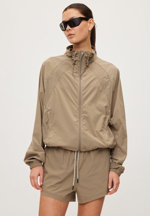 Beige Windbreaker-Jacke mit hohem Kragen, Reißverschluss vorne und elastischen Bündchen, kombiniert mit passenden Shorts und sportlichen Sonnenbrillen.