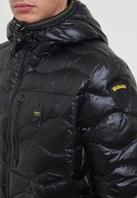 Giacca puffer nera trapuntata con finitura lucida, cappuccio regolabile, zip frontale e logo a patch sulla spalla. Presenta dettagli gialli.