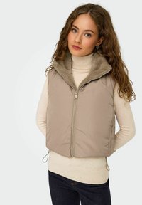 Beige puffer vest met een zachte, nep-bont kraag, beschikt over een ritssluiting aan de voorkant en verstelbare trekkoorden. Draagt over een licht truitje.