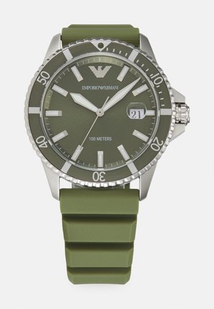 Montre avec bracelet en silicone vert, boîtier rond en argent, grands index horaires, affichage de la date et résistance à l'eau jusqu'à 100 mètres.
