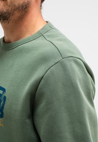 Groene katoenen sweatshirt met een geribbelde ronde hals, schoudernaden en een blauwe grafische print met tekst aan de voorkant. Zachte textuur.