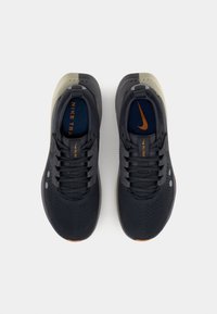 Chaussures de sport noires avec une tige en maille, texture perforée, accents marron clair, logo orange et semelle amortie. Dotées d'un lacet traditionnel.