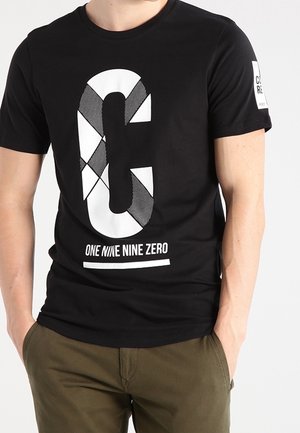 T-shirt noir avec une grande lettre stylisée "C" et le texte "ONE NINE NINE ZERO" porté par une personne les mains dans les poches d'un pantalon vert olive.