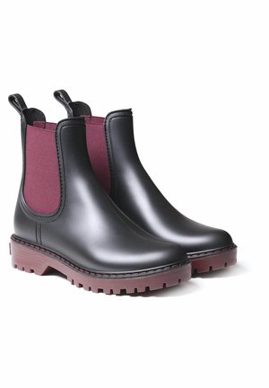 Paire de bottines Chelsea en caoutchouc noir avec panneaux latéraux élastiques rouge foncé et semelles à crampons assorties sur fond blanc.