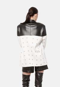 Czarny i biały płaszcz z skórzaną akcentem i wzorzystą dolną częścią. Projekt zawiera małe logo na białej tkaninie.