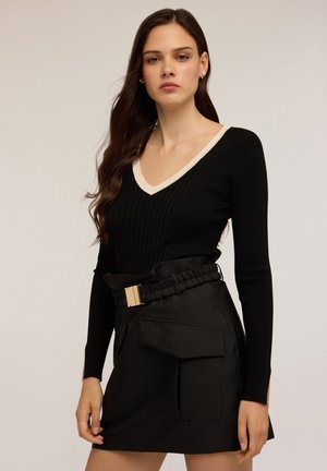 Donna con capelli lunghi e scuri, vestita con un maglione a coste nero con scollatura a V e rifiniture beige, e una gonna nera alta in vita con una grande tasca frontale e cintura.