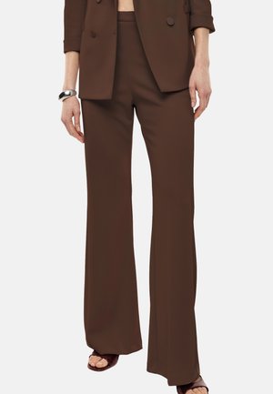 Donna che indossa pantaloni marroni a gamba larga e blazer abbinato, accessoriati con un braccialetto d'argento e sandali marroni con punta aperta.