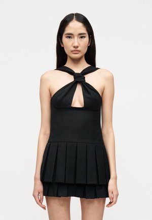 Νεαρή γυναίκα με μακριά σκούρα μαλλιά φοράει μαύρο αμάνικο φόρεμα με δέσιμο στον λαιμό και πολυστρωματιστή πλισέ φούστα, στέκεται μπροστά σε απλό λευκό φόντο.