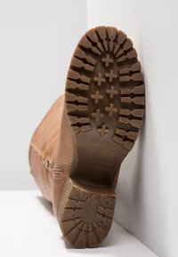 Semelle de botte marron avec une bande de roulement profonde et des motifs en forme de croix, inclinée contre un mur blanc et une surface.