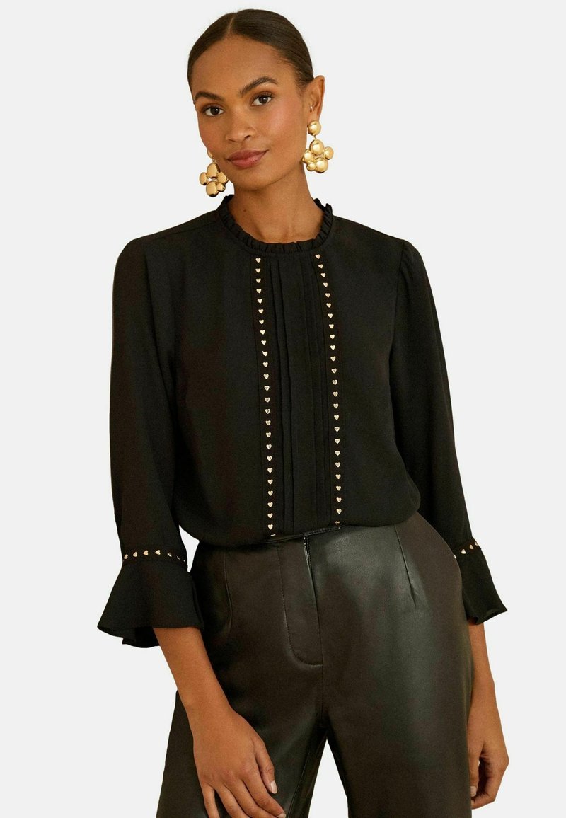 Juoda palaidinė su raukiniais ant apykaklės, ilgomis rankovėmis ir aukso spalvos širdies formos smeigtukais priekyje ir ant rankogalų. Derinama su juodomis odinėmis kelnėmis.