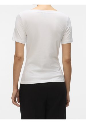 Femme portant un t-shirt blanc uni à manches courtes et un pantalon noir, vue de dos sur un fond uni.