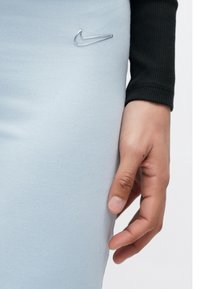 Svetlomodré legíny z pružnej látky s hladkým povrchom, s kovovým strieborným logom Nike na ľavej hornej strane. Viditeľné čierne tričko s dlhými rukávmi.