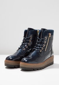 Stivali alla caviglia in pelle verniciata blu navy con lacci sul davanti, zip laterale, minuteria dorata e suole in piattaforma marrone ruvido. Finitura lucida con dettagli in camoscio.