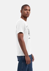 Weißes Baumwoll-T-Shirt mit einem grafischen Design auf der Vorderseite, kurzen Ärmeln und einem lässigen Schnitt. Getragen mit blauen Jeans.