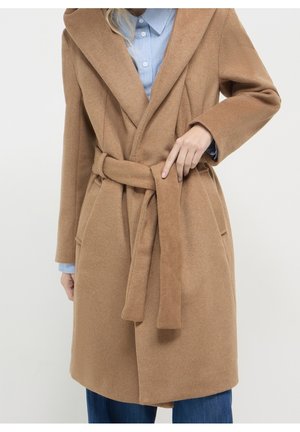 Frau trägt einen wollenen Mantel in Kamelbraun mit Gürtel über einem blau gestreiften Hemd und blauen Jeans, sie justiert den Mantelgürtel mit der rechten Hand.