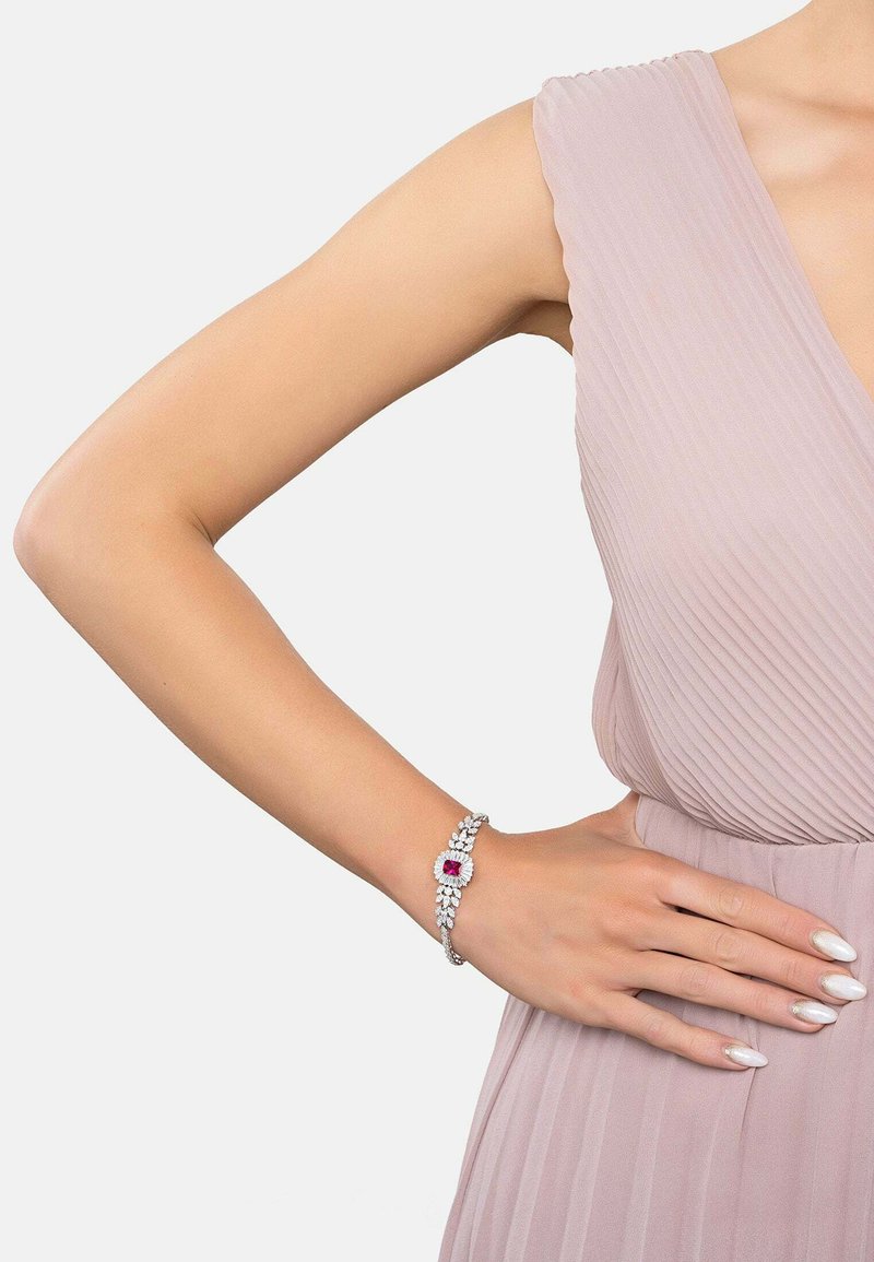 Silver bracelet featuring a central red stone, surrounded by clear rhinestones, set against a light pink pleated dress with a deep neckline.