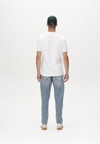 Bela kratka majica s kratkimi rokavi in okroglim izrezom, v kombinaciji s svetloblujimi jeans hlaci. Model nosi temnozeleno kapo in bele superge.