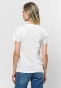 Balta kokvilnas T-kreklu ar īsām piedurknēm, taisnu apakšmalu un apaļu kakla izgriezumu, valkāta kopā ar ziliem džinsiem, skatīta no aizmugures.