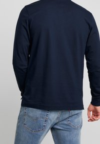 Muž v tmavě modré košili s dlouhým rukávem a světlých džínech, zobrazený zezadu na jednobarevném pozadí.