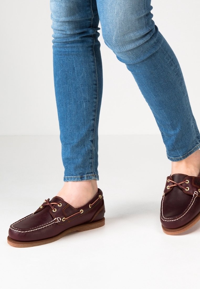 Personne portant un jean bleu et des chaussures bateau en cuir marron avec des coutures blanches et des semelles en bois sur un sol blanc.