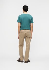 Νεαρός άνδρας με την πλάτη, φοράει τιρκουάζ μπλουζάκι, μπεζ παντελόνι και σκούρα καφέ παπούτσια, μπροστά σε απλό λευκό φόντο.